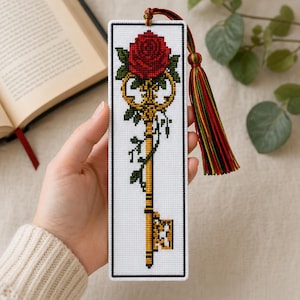 May include: A rectangular cross-stitch bookmark on white fabric featuring a gold skeleton key entwined with green vines and topped with a vibrant red rose. The bookmark is finished with a multi-coloured tassel in shades of red, green, and gold.