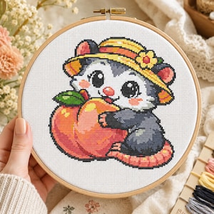May include: A circular wooden embroidery hoop containing a cross-stitch pattern of a grey and white opossum wearing a yellow sun hat with a small flower, holding a large orange peach against a white fabric background with a visible grid texture.