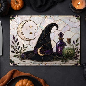 Puede incluir: Una tabla de cortar rectangular de vidrio con una ilustración estilo vidriera que muestra un sombrero de bruja negro con una banda morada y una luna creciente, una luna creciente en el cielo y dos frascos de pociones sobre un fondo iridiscente.