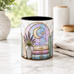 May include: A cylindrical ceramic mug with a black interior and rim. The exterior features a stained-glass style illustration of a crescent moon, a star, a vase of purple lavender, and a stack of books in shades of white, pink, and blue.