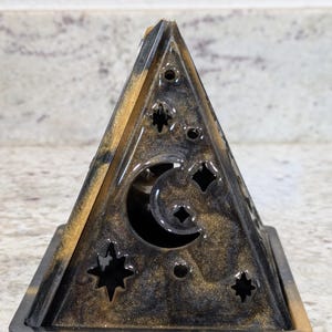 May include: A triangular pyramid-shaped object made of black and gold swirled resin. The structure features cutouts in the shapes of a crescent moon and various stars on its front face, set upon a square base with matching black and gold colors.