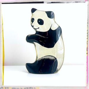 Puede incluir: Una figurita de panda blanco y negro hecha de vidrio. El panda está de pie sobre sus patas traseras con las patas delanteras levantadas. La figurita es de vidrio transparente con detalles pintados en negro.