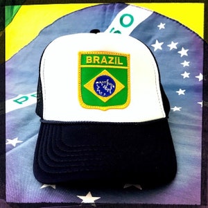 以下が含まれることがあります： 白と黒のトラッカーハットに、緑、黄色、青の刺繍が施された「BRAZIL」と書かれたパッチが付いています。帽子は、星がついた青と白の布の上に置かれています。
