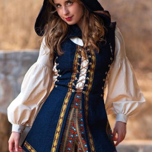 Op de afbeelding: Een donkerblauw vest met capuchon, voorzien van ingewikkelde gouden biezen en vetersluiting aan de zijkant, gedragen over een crèmekleurig shirt met lange, volumineuze mouwen en een broek met rode, blauwe en gouden verticale strepen.