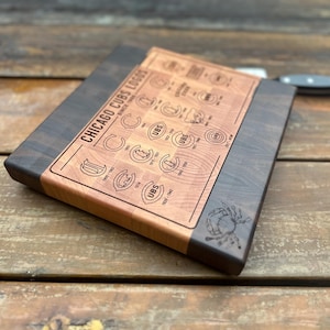 May include: A wooden cutting board featuring the Chicago Cubs logos. The board has a dark wood border and a lighter wood center with engraved logos and text. A knife rests on the wooden surface.