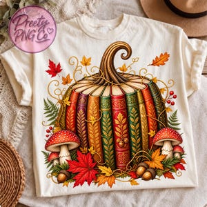 Può includere: Una t-shirt color crema con la grafica di una zucca composta da libri verticali e colorati in stile vintage. Il design include foglie autunnali rosse e arancioni, funghi dal cappello rosso e ghiande marroni attorno alla base.