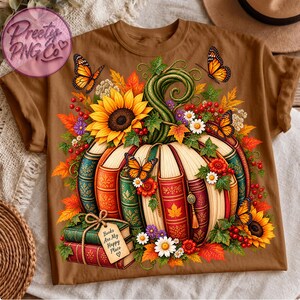 Op de afbeelding: Een bruin t-shirt met een afbeelding van een pompoen gemaakt van gestapelde boeken, versierd met zonnebloemen, herfstbladeren, bessen en monarchvlinders. Een kleine stapel boeken met een label "Books Are My Happy Place" staat onderaan.