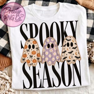 May include: A white short-sleeve t-shirt featuring the black text SPOOKY SEASON and three ghost illustrations. The first ghost has an orange pumpkin and black bow pattern, the second has a purple daisy pattern, and the third has a black bat and moon pattern.