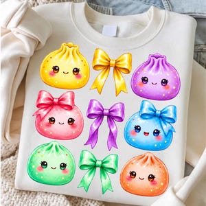 May include: A cream-colored crewneck sweatshirt featuring a graphic print of six colorful, smiling dumpling characters and three decorative bows. The dumplings are yellow, purple, pink, blue, green, and orange, each with sparkling star accents.