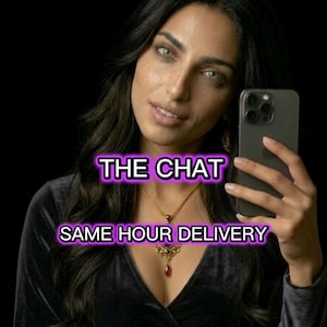 May include: A woman with long dark hair and green eyes holds a black smartphone. She wears a black velvet top and a gold necklace with a red pendant. The text "THE CHAT" and "SAME HOUR DELIVERY" are displayed in a purple font.