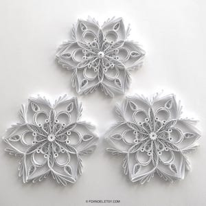 May include: Three white paper quilling snowflakes featuring intricate, symmetrical designs made from coiled and shaped paper strips. Each snowflake displays a complex pattern of loops, teardrops, and scrolls arranged in a radial, star-like formation.