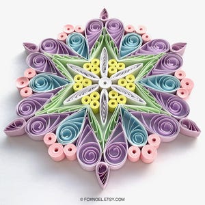 May include: A paper quilling art piece in a star or snowflake shape, featuring intricate coiled paper strips in pastel shades of lavender, light blue, mint green, pale yellow, and soft pink. The text FOXNOEL.ETSY.COM is printed at the bottom.