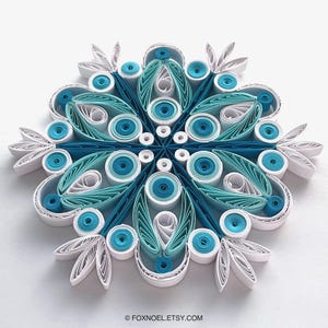 May include: A paper quilling snowflake ornament featuring intricate concentric circles and teardrop shapes in shades of white, light blue, and dark teal. The design is symmetrical with layered paper strips forming a complex, star-like pattern.