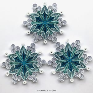 May include: Three paper quilling snowflake ornaments in shades of teal, light blue, and white. Each snowflake features intricate coiled paper designs with heart-shaped petals and circular accents. The text FOXNOEL.ETSY.COM is printed at the bottom.