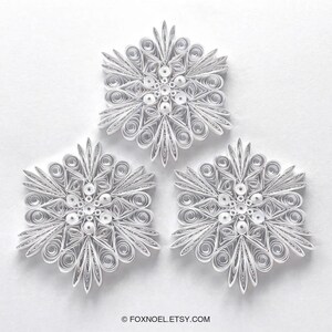 May include: Three white paper quilling snowflakes featuring intricate coiled and looped designs arranged in a symmetrical pattern. Each snowflake is constructed from thin paper strips with a central circular element and the text FOXNOEL.ETSY.COM.