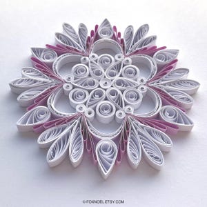 May include: A circular paper quilling ornament featuring intricate white and light pink coiled paper strips arranged in a symmetrical floral pattern. The design consists of numerous teardrop and spiral shapes radiating from the center, with the text FOXNOEL.ETSY.COM printed at the bottom.