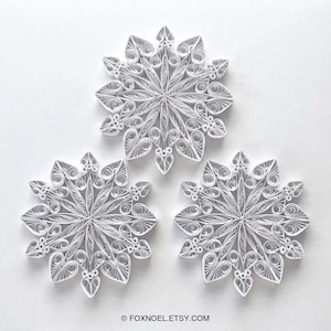 May include: Three white paper quilling snowflakes featuring intricate coiled and looped designs. Each snowflake is composed of delicate paper strips arranged in a symmetrical star pattern with heart-shaped and circular elements. Text reads FOXNOEL.ETSY.COM.