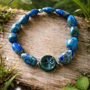 Könnte beinhalten: Ein Armband mit blauen und silberfarbenen Perlen und einem türkisfarbenen Libellen-Charm. Das Armband wird auf Holz präsentiert, mit einer Libelle im Hintergrund. Die Perlen haben einen Durchmesser von etwa 1 cm.