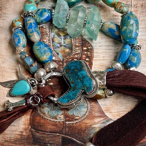 May include: A turquoise and silver necklace with a western theme. The necklace features a turquoise boot pendant, turquoise and blue stone beads, and clear crystal accents. Brown suede straps and silver accents complete the design.