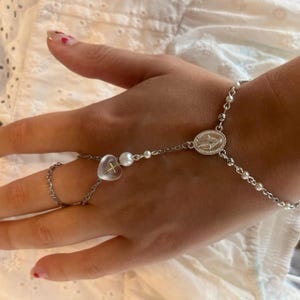 Könnte beinhalten: Eine silberfarbene Handkette mit einer feinen Metallkette, einer kleinen, durchsichtigen, herzförmigen Perle mit einem goldenen Kreuz im Inneren, einer weißen perlenartigen Kugel und einer ovalen Medaille in der Mitte mit einer Figur.