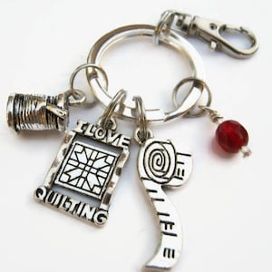 May include: Silver keychain with charms including a spool of thread, a quilt block, a measuring tape, and a red bead. The charm with the quilt block says "I love quilting".