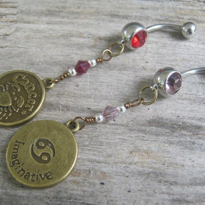May include: Two silver belly button rings with charms. The charms are round and feature the zodiac sign Cancer. One charm says "Cancer" and the other says "Imaginative".