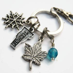 May include: Silver keychain with charms including a snowflake, a maple leaf, a blue glass bead, and a charm shaped like the state of Vermont with the word "VERMONT" written on it.