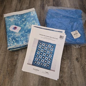 May include: A Twinkling Stars Teal Quilt Kit by Monique Dillard featuring Boundless Blenders Botanical fabric. The kit includes a printed pattern sheet with a finished quilt image, a package of blue floral fabric, and a package of solid blue fabric.