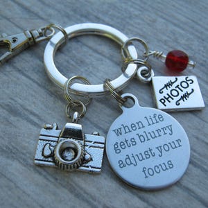 May include: Silver keychain with a camera charm, a round charm with the text "when life gets blurry adjust your focus", and a charm with the text "PHOTOS".