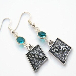 May include: A pair of silver dangle earrings featuring a turquoise blue crystal and a silver charm shaped like the state of New Mexico with the text "New Mexico" on it.