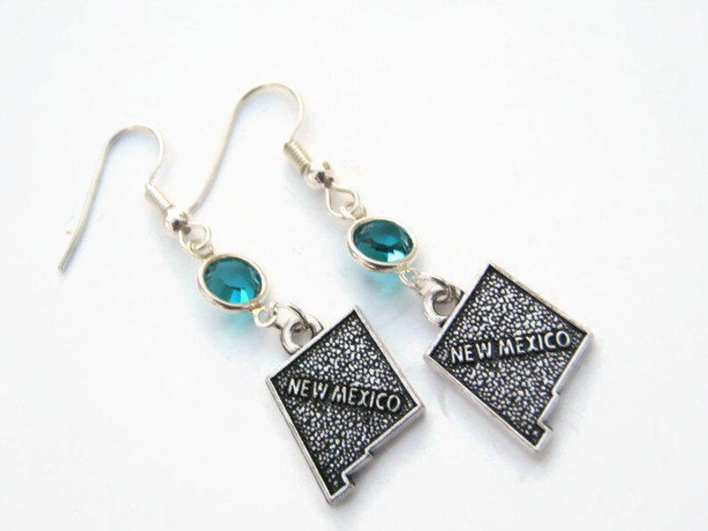 May include: A pair of silver dangle earrings featuring a turquoise blue crystal and a silver charm shaped like the state of New Mexico with the text "New Mexico" on it.