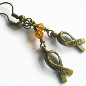 Puede incluir: Un par de pendientes de bronce con una peque&ntilde;a cuenta de cristal &aacute;mbar y un dije de cinta de bronce con la palabra "HOPE" grabada.