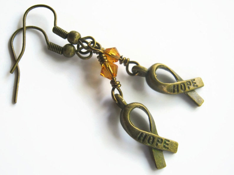 Puede incluir: Un par de pendientes de bronce con una peque&ntilde;a cuenta de cristal &aacute;mbar y un dije de cinta de bronce con la palabra "HOPE" grabada.