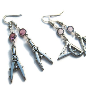 Puede incluir: Pendientes colgantes plateados con diseño de compás y transportador. Cada pendiente tiene dos gemas rosas y un cierre de gancho. Los pendientes están sobre un fondo blanco.