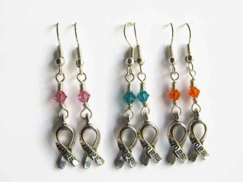 May include: Three pairs of silver earrings with a silver ribbon charm that says "HOPE". Each pair has a different colored crystal bead: pink, teal, and orange.