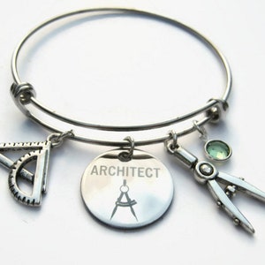 May include: A silver bangle bracelet with charms. The charms include a protractor and compass, a round charm with the word "ARCHITECT" and a compass design, and a green gemstone.
