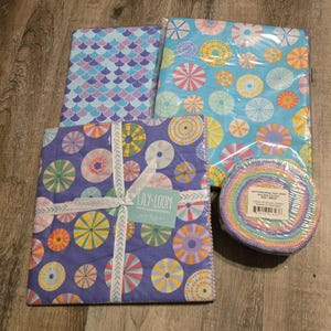 May include: Four fabric bundles in various patterns. One bundle features a mermaid scale design in shades of blue and purple. Another has a blue background with colorful circular designs. A third bundle has a purple background with colorful circular designs. A fourth bundle is a roll of fabric strips.