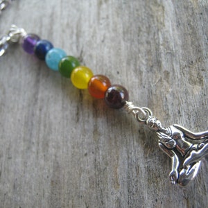 May include: A silver chain necklace with a silver charm of a person sitting in a yoga pose. The necklace has seven beads in rainbow colors: purple, blue, teal, green, yellow, orange, and red.
