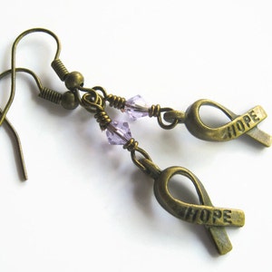 Puede incluir: Un par de pendientes de bronce con una cuenta de cristal morado y un dije de cinta de bronce con la palabra "HOPE" grabada.