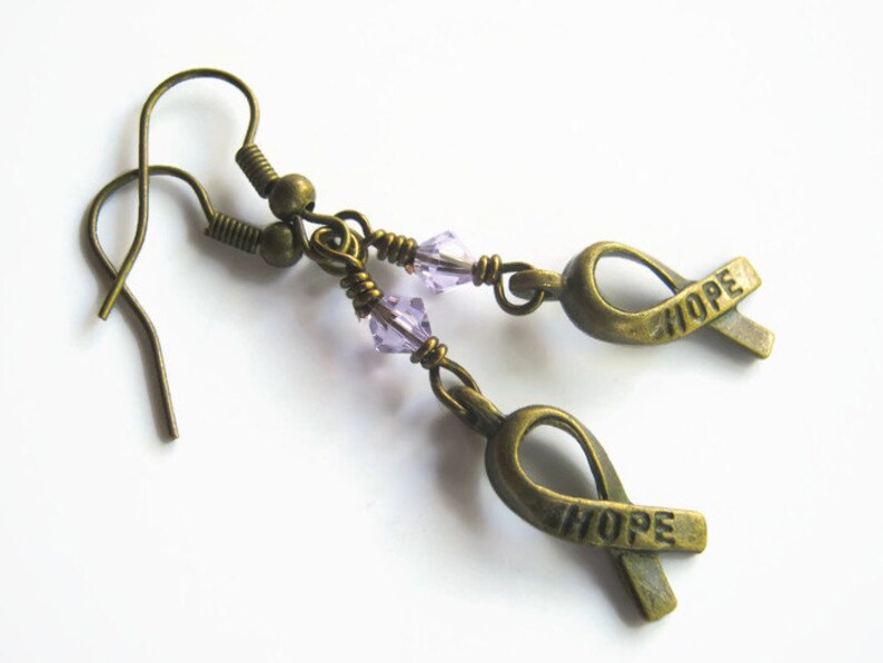 Puede incluir: Un par de pendientes de bronce con una cuenta de cristal morado y un dije de cinta de bronce con la palabra "HOPE" grabada.
