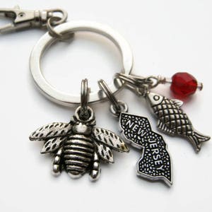 Puede incluir: Llavero de plata con un dije de abeja, un dije del estado de Nueva Jersey, un dije de pez y una cuenta roja.