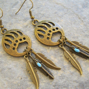 May include: A pair of bronze earrings with a bear paw print design and dangling feathers with turquoise accents.