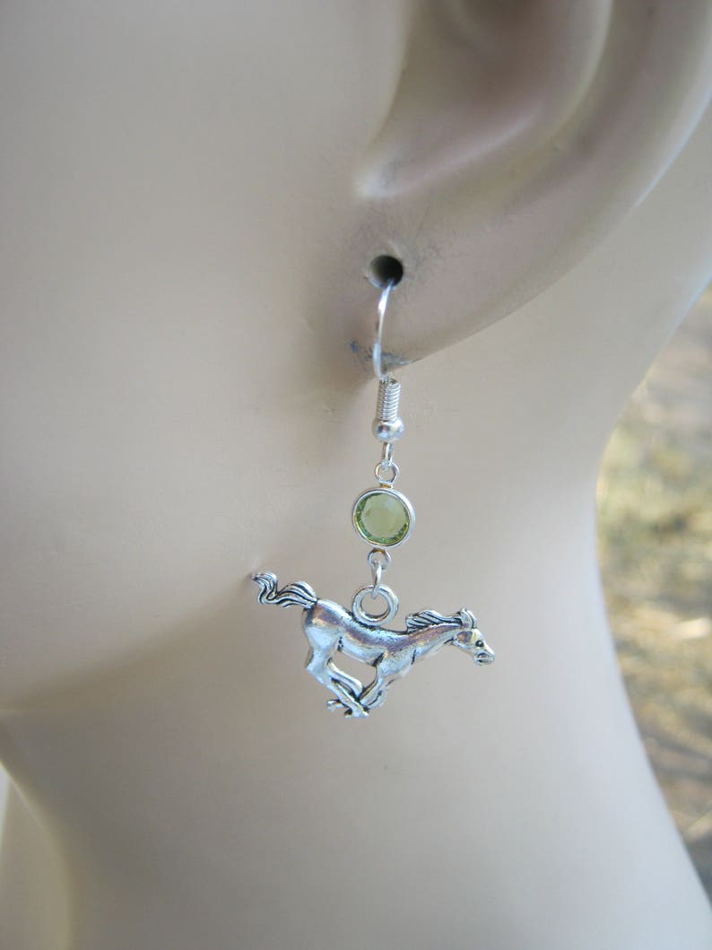 May include: Silver dangle earrings with a green gemstone and a silver horse charm.