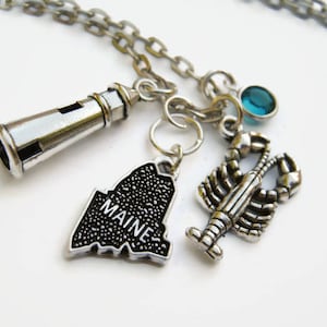 May include: A silver necklace with charms representing Maine. The charms include a lighthouse, a lobster, and the outline of the state with the word "MAINE" printed on it. There is also a small blue gemstone charm.