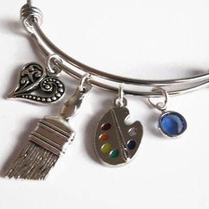 May include: Silver charm bracelet with a heart, paintbrush, paint palette, and blue crystal charm. The bracelet is expandable and has a silver finish.