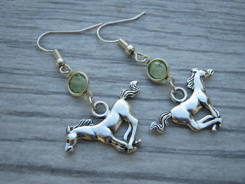May include: Silver horse earrings with green gemstone accents. The horses are in a running pose.