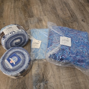 May include: Two fabric jelly rolls featuring blue and white swirling patterns and two folded fabric squares in clear plastic packaging, each with a white product label containing text and a barcode, arranged on a wood-grain surface.