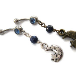 May include: Two belly button rings with a celestial theme. Each ring has a curved barbell with a blue gem. One features a silver crescent moon charm with stars, the other a bronze moon charm.