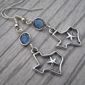 May include: Silver earrings featuring a blue crystal and a cutout of the state of Texas with a star inside.