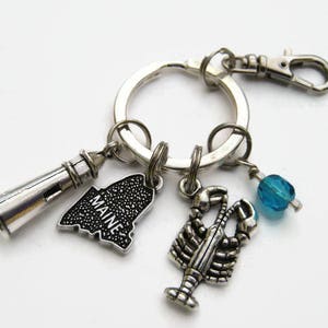 May include: Silver keyring with a lobster, lighthouse, and a charm featuring the state of Maine with the word "MAINE" on it. The keyring also has a blue glass bead.